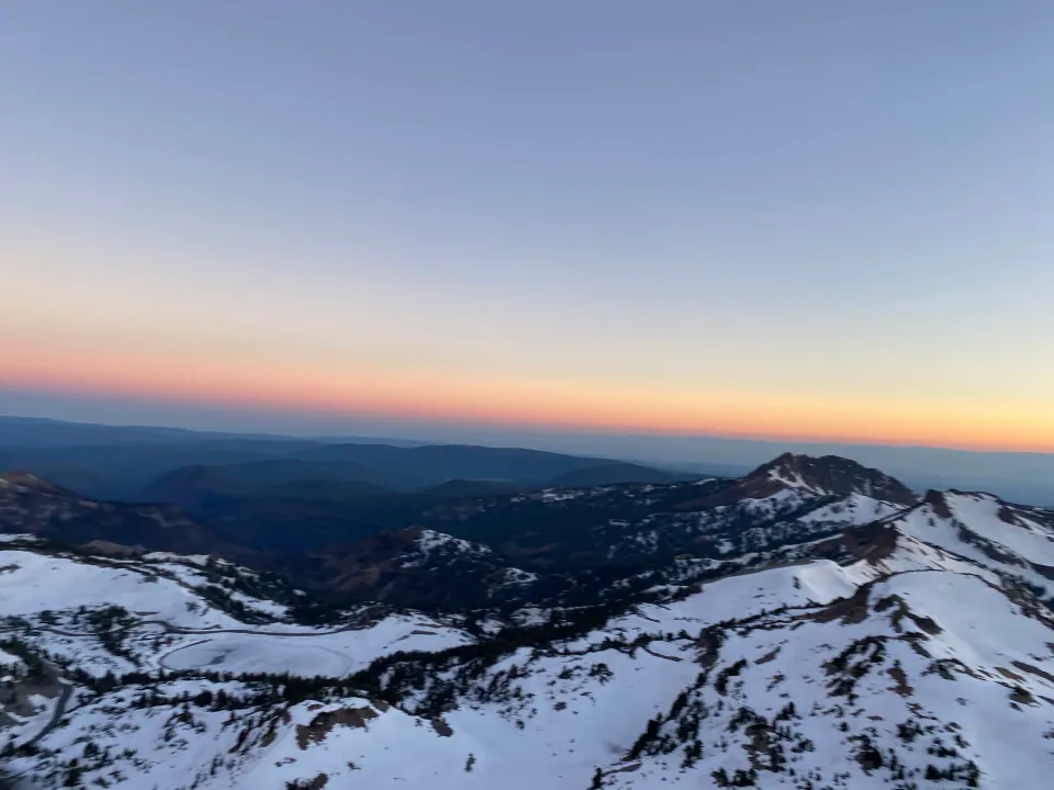 Snowy peaks at dawn from summit, orange glow along horizon