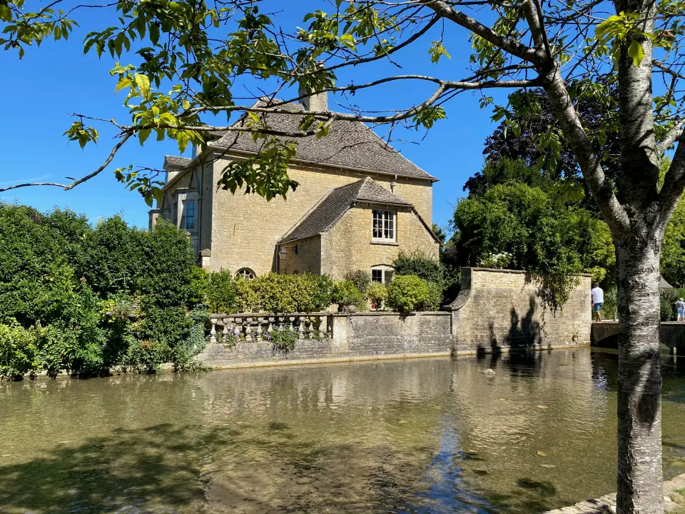 Cotswold stone cottage reflected in river, summer trees