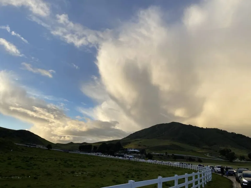 Storm cloud building over a green hill above a white ranch fence